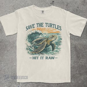 May include: Cream-colored t-shirt featuring a sea turtle swimming in the ocean, with a wave and sunset graphic. The text "SAVE THE TURTLES" and "- HIT IT RAW -" are printed above and below the turtle illustration.