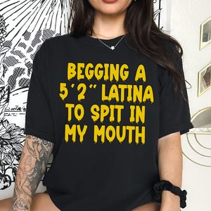 May include: Black t-shirt with yellow text that reads "BEGGING A 5'2" LATINA TO SPIT IN MY MOUTH". The text is in a bold, dripping font. The shirt is a crew neck style.