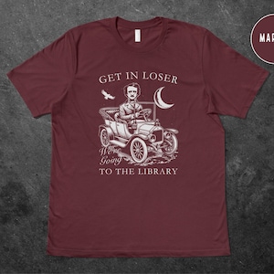 May include: A maroon t-shirt with a white graphic of Edgar Allan Poe driving a vintage car with the text "Get in loser, we're going to the library".