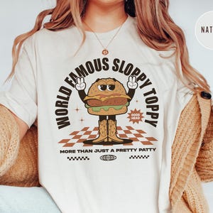 May include: White t-shirt featuring a cartoon burger wearing cowboy boots and the text "World Famous Sloppy Toppy" and "More Than Just A Pretty Patty".