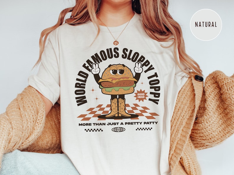 May include: White t-shirt featuring a cartoon burger wearing cowboy boots and the text "World Famous Sloppy Toppy" and "More Than Just A Pretty Patty".