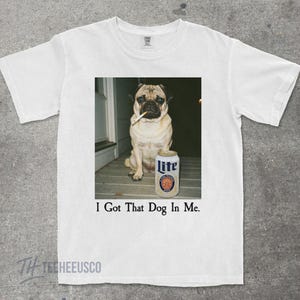Pu&ograve; includere: T-shirt bianco con un carlino che fuma una sigaretta, accanto a una lattina di birra Lite. Il testo "I Got That Dog In Me" &egrave; stampato sotto l'immagine. La maglietta ha uno scollo tondo.