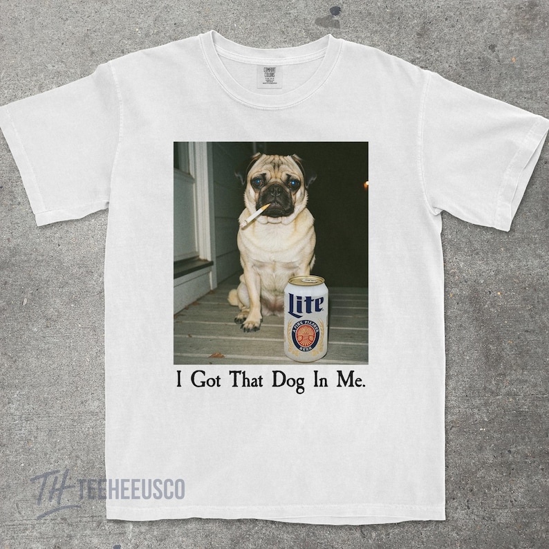 Pu&ograve; includere: T-shirt bianco con un carlino che fuma una sigaretta, accanto a una lattina di birra Lite. Il testo "I Got That Dog In Me" &egrave; stampato sotto l'immagine. La maglietta ha uno scollo tondo.