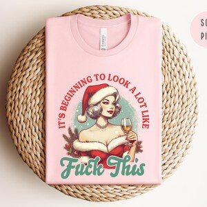 Pu&ograve; includere: T-shirt rosa chiaro con grafica di una donna con cappello da Babbo Natale che tiene un bicchiere di vino. Il testo recita "It's beginning to look a lot like Fuck This" in rosso e turchese. La maglietta &egrave; piegata su una superficie intrecciata.