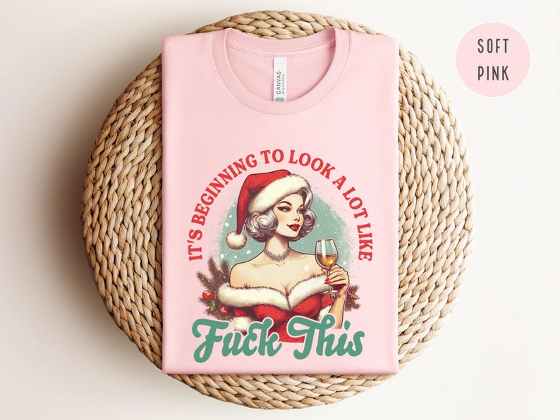 Pu&ograve; includere: T-shirt rosa chiaro con grafica di una donna con cappello da Babbo Natale che tiene un bicchiere di vino. Il testo recita "It's beginning to look a lot like Fuck This" in rosso e turchese. La maglietta &egrave; piegata su una superficie intrecciata.