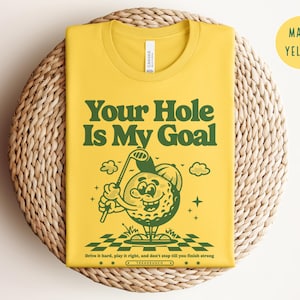 Może przedstawiać: Ż&oacute;łta koszulka z zieloną kresk&oacute;wkową postacią piłki golfowej trzymającą kij golfowy. Tekst "Your Hole Is My Goal" jest nadrukowany na koszulce zielonymi literami. Tekst "Drive it hard, play it right, and don't stop till you finish strong" jest nadrukowany mniejszymi zielonymi literami pod kresk&oacute;wką. Koszulka leży na plecionym słomianym podłożu.