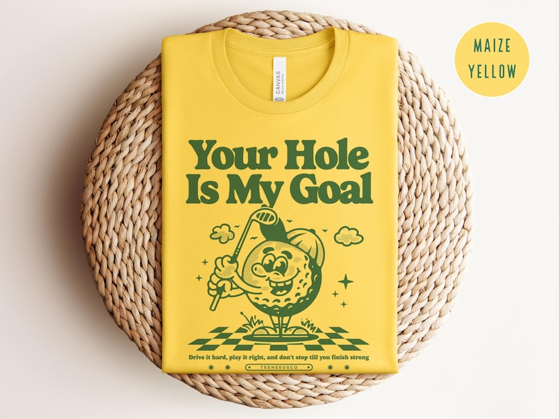 Może przedstawiać: Ż&oacute;łta koszulka z zieloną kresk&oacute;wkową postacią piłki golfowej trzymającą kij golfowy. Tekst "Your Hole Is My Goal" jest nadrukowany na koszulce zielonymi literami. Tekst "Drive it hard, play it right, and don't stop till you finish strong" jest nadrukowany mniejszymi zielonymi literami pod kresk&oacute;wką. Koszulka leży na plecionym słomianym podłożu.