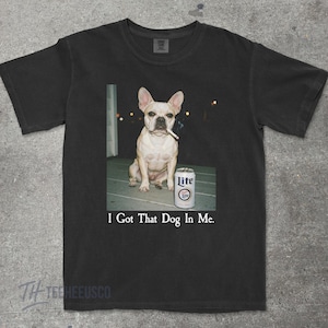 May include: Black t-shirt featuring a French Bulldog with a cigarette in its mouth, sitting next to a can of Lite beer. The text "I Got That Dog In Me" is printed below the image.