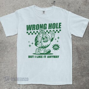 May include: Light blue t-shirt with a green graphic of a golf ball character holding a golf club and giving a peace sign. The text reads "WRONG HOLE" above the character and "BUT I LIKE IT ANYWAY" below.