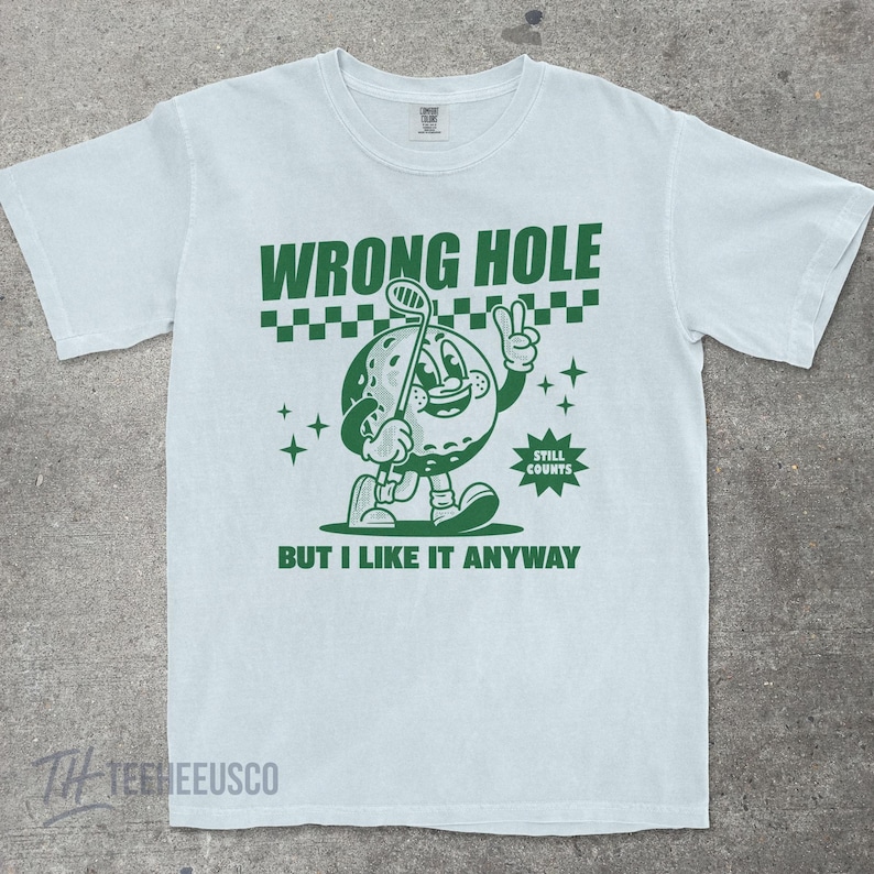 May include: Light blue t-shirt with a green graphic of a golf ball character holding a golf club and giving a peace sign. The text reads "WRONG HOLE" above the character and "BUT I LIKE IT ANYWAY" below.