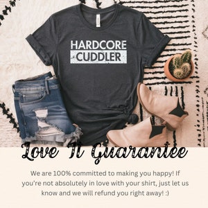 Puede incluir: Una camiseta gris oscuro con el texto "HARDCORE CUDDLER" impreso en blanco. La camiseta est&aacute; colocada sobre una alfombra de rayas blancas y negras con un par de pantalones cortos de mezclilla desgastados, un par de botines de tobillo color canela y una peque&ntilde;a planta suculenta en maceta.