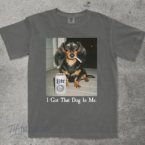 May include: Dark gray t-shirt featuring a dachshund with a cigarette in its mouth and a can of Lite beer. The text "I Got That Dog In Me" is printed below the image. The shirt is made of a soft material.