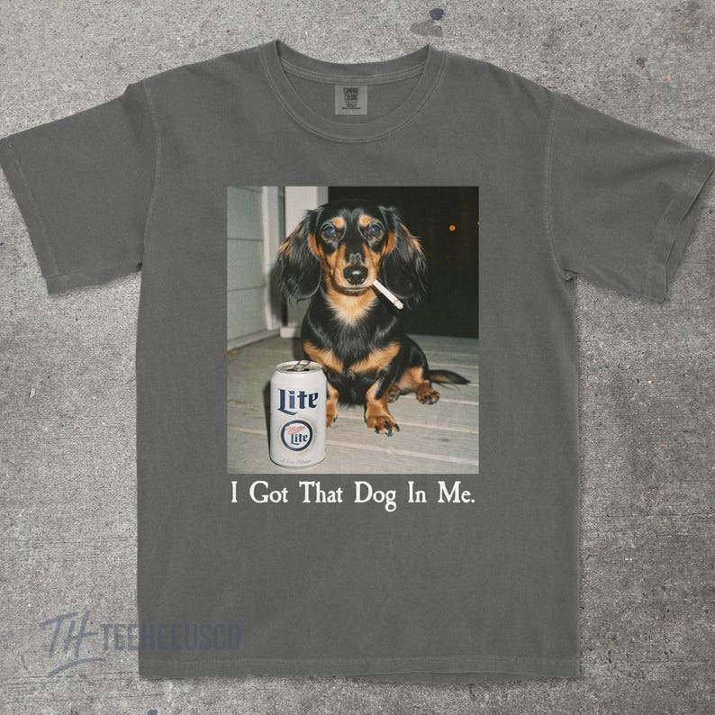 May include: Dark gray t-shirt featuring a dachshund with a cigarette in its mouth and a can of Lite beer. The text "I Got That Dog In Me" is printed below the image. The shirt is made of a soft material.