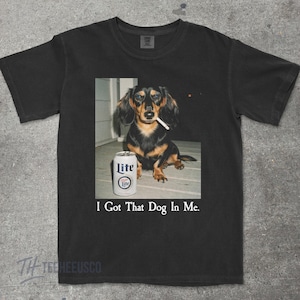 May include: Black t-shirt featuring a photo of a dachshund with a cigarette in its mouth, next to a can of Lite beer. The text "I Got That Dog In Me" is printed below the image. The shirt is made of a soft material.
