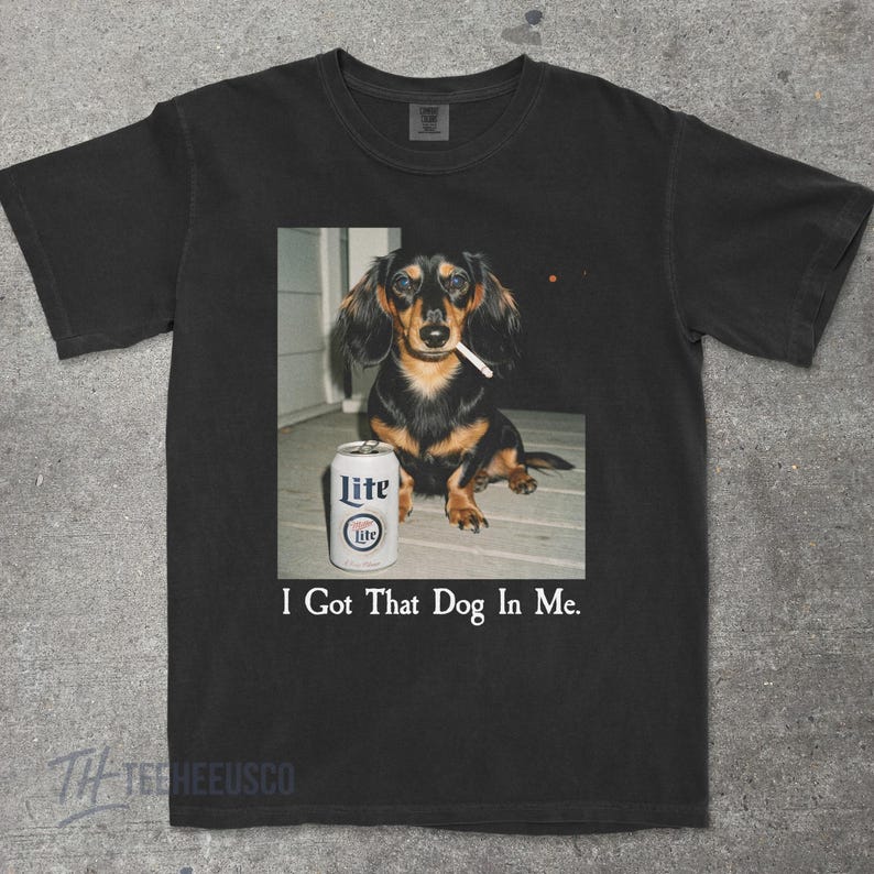 May include: Black t-shirt featuring a photo of a dachshund with a cigarette in its mouth, next to a can of Lite beer. The text "I Got That Dog In Me" is printed below the image. The shirt is made of a soft material.