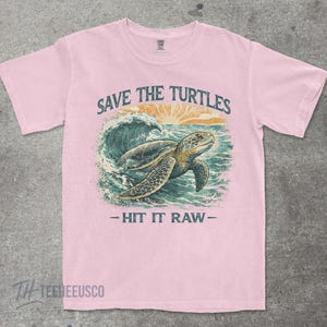 May include: Light pink t-shirt with a graphic of a sea turtle swimming in the ocean. The text "SAVE THE TURTLES" is above the turtle, and "- HIT IT RAW -" is below. The shirt has a crew neck and short sleeves.