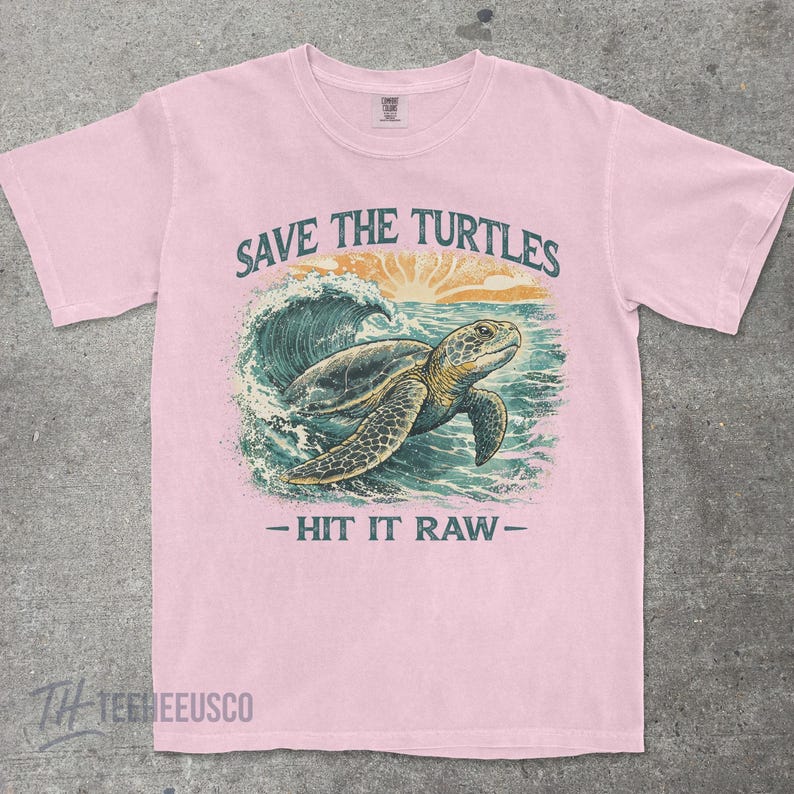 May include: Light pink t-shirt with a graphic of a sea turtle swimming in the ocean. The text "SAVE THE TURTLES" is above the turtle, and "- HIT IT RAW -" is below. The shirt has a crew neck and short sleeves.