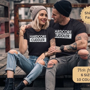 Puede incluir: Una pareja vistiendo camisetas negras con texto blanco que dice "HARDCORE CUDDLER". La mujer lleva una boina gris y pantalones vaqueros rotos. El hombre tiene tatuajes en los brazos y lleva pantalones vaqueros negros.