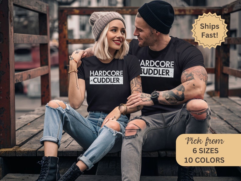 Puede incluir: Una pareja vistiendo camisetas negras con texto blanco que dice "HARDCORE CUDDLER". La mujer lleva una boina gris y pantalones vaqueros rotos. El hombre tiene tatuajes en los brazos y lleva pantalones vaqueros negros.