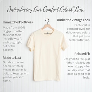 K&ouml;nnte beinhalten: Cremefarbenes T-Shirt, das an einem Holzb&uuml;gel h&auml;ngt. Das Shirt geh&ouml;rt zur "Comfort Colors Line" und enth&auml;lt Text, der seine Weichheit, Haltbarkeit und entspannte Passform beschreibt. Das Shirt besteht aus Ringspun-Baumwolle.