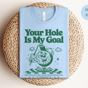 Może przedstawiać: Jasnoniebieska koszulka z zielonym, kreskowym bohaterem w postaci piłki golfowej trzymającym kij golfowy. Na koszulce nadrukowany jest zielony tekst "Your Hole Is My Goal". Pod bohaterem nadrukowany jest czarny tekst "Drive it hard, play it right, and don't stop till you finish strong".