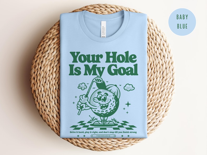 Może przedstawiać: Jasnoniebieska koszulka z zielonym, kreskowym bohaterem w postaci piłki golfowej trzymającym kij golfowy. Na koszulce nadrukowany jest zielony tekst "Your Hole Is My Goal". Pod bohaterem nadrukowany jest czarny tekst "Drive it hard, play it right, and don't stop till you finish strong".