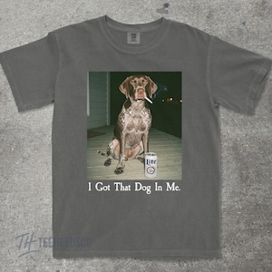 K&ouml;nnte beinhalten: Graues T-Shirt mit einem Aufdruck eines Hundes, der eine Zigarette raucht, neben einer Lite-Bierdose. Der Text "I Got That Dog In Me" ist unter dem Bild aufgedruckt. Das T-Shirt hat einen Rundhalsausschnitt und kurze &Auml;rmel. Der Hund ist braun und wei&szlig;.