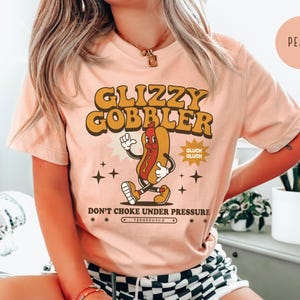 K&ouml;nnte beinhalten: Pfirsichfarbenes T-Shirt mit einem Retro-Grafik eines laufenden Hotdog-Charakters. Der Text "GLIZZY GOBBLER" befindet sich &uuml;ber dem Hotdog, mit "DON'T CHOKE UNDER PRESSURE" darunter. Das Shirt hat eine Vintage-&Auml;sthetik.