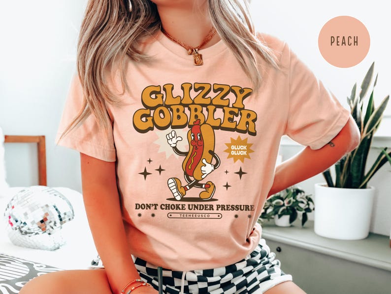 K&ouml;nnte beinhalten: Pfirsichfarbenes T-Shirt mit einem Retro-Grafik eines laufenden Hotdog-Charakters. Der Text "GLIZZY GOBBLER" befindet sich &uuml;ber dem Hotdog, mit "DON'T CHOKE UNDER PRESSURE" darunter. Das Shirt hat eine Vintage-&Auml;sthetik.