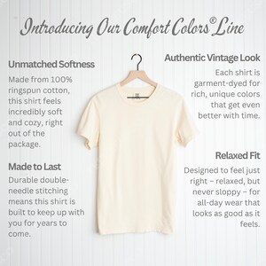May include: A cream-colored t-shirt hangs on a wooden hanger against a white background. The shirt is part of the Comfort Colors line, made from ringspun cotton. Text on the left describes the shirt's softness and durability. Text on the right highlights its vintage look and relaxed fit.