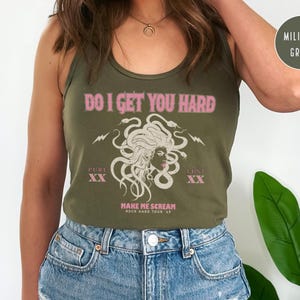 May include: An olive green vest top with the pink text "DO I GET YOU HARD". The graphic features a Medusa illustration with the words "PURE XX" and "LUST XX". The text "MAKE ME SCREAM ROCK HARD TOUR 69" is also present.