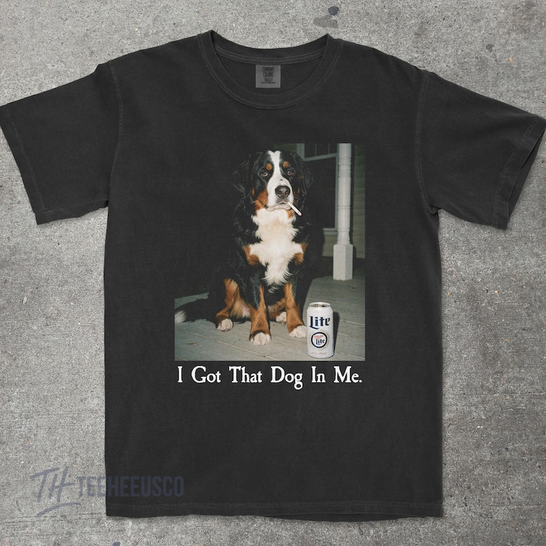 May include: Black t-shirt featuring a Bernese Mountain Dog with a cigarette in its mouth, sitting next to a can of Lite beer. The text "I Got That Dog In Me" is printed below the image. The shirt has a crew neck.