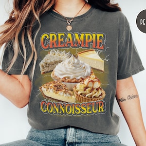May include: A gray t-shirt with the text "CREAMPIE CONNOISSEUR" in orange and yellow letters. The shirt features a graphic of various cream pies, including a cheesecake, a pecan pie, and a lemon meringue pie.