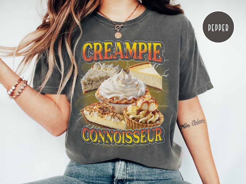 May include: A gray t-shirt with the text "CREAMPIE CONNOISSEUR" in orange and yellow letters. The shirt features a graphic of various cream pies, including a cheesecake, a pecan pie, and a lemon meringue pie.
