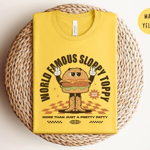 May include: A yellow t-shirt featuring a cartoon burger wearing a chef's hat and holding a sign that says "Good Head". The text on the shirt reads "World Famous Sloppy Toppy More Than Just A Pretty Patty".