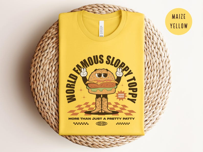 May include: A yellow t-shirt featuring a cartoon burger wearing a chef's hat and holding a sign that says "Good Head". The text on the shirt reads "World Famous Sloppy Toppy More Than Just A Pretty Patty".
