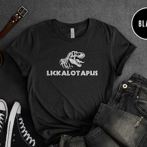 Może przedstawiać: Czarny T-shirt z białym nadrukiem szkieletu dinozaura i tekstem "LICKALOTAPUS".