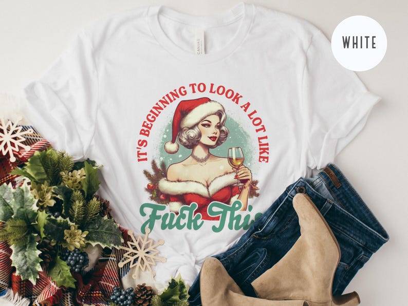 Pu&ograve; includere: T-shirt bianco con un'illustrazione retr&ograve; di una donna con un cappello da Babbo Natale che tiene in mano un bicchiere di vino. Il testo recita "It's beginning to look a lot like Fuck This". La maglietta &egrave; abbinata a jeans blu e stivali color cammello.