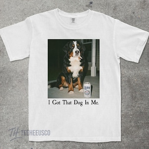 May include: White t-shirt featuring a graphic of a Bernese Mountain Dog with a cigarette in its mouth, next to a can of Lite beer. The text "I Got That Dog In Me." is printed below the image.