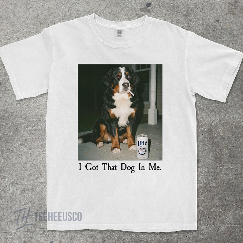 May include: White t-shirt featuring a graphic of a Bernese Mountain Dog with a cigarette in its mouth, next to a can of Lite beer. The text "I Got That Dog In Me." is printed below the image.