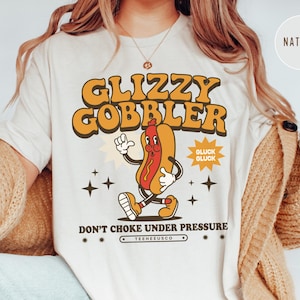 K&ouml;nnte beinhalten: Cremefarbenes T-Shirt mit Retro-Grafik. Das Design zeigt einen Cartoon-Hotdog-Charakter mit dem Text "GLIZZY GOBBLER" in Orange und Gelb. Darunter steht "DON'T CHOKE UNDER PRESSURE".