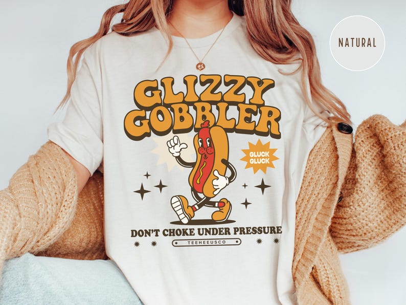 K&ouml;nnte beinhalten: Cremefarbenes T-Shirt mit Retro-Grafik. Das Design zeigt einen Cartoon-Hotdog-Charakter mit dem Text "GLIZZY GOBBLER" in Orange und Gelb. Darunter steht "DON'T CHOKE UNDER PRESSURE".