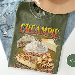 May include: Green t-shirt with the text "CREAMPIE CONNOISSEUR" in yellow and orange letters. The shirt features a graphic of four different types of pies, including a cheesecake, a cream pie, and a pecan pie.