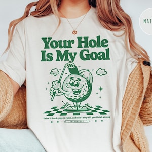 Może przedstawiać: Biały t-shirt z zielonym nadrukiem przedstawiającym kresk&oacute;wkową piłkę golfową trzymającą kij golfowy. Tekst "Your Hole Is My Goal" jest nadrukowany na zielono nad grafiką. Tekst "Drive it hard, play it right, and don't stop till you finish strong" jest nadrukowany na zielono pod grafiką.