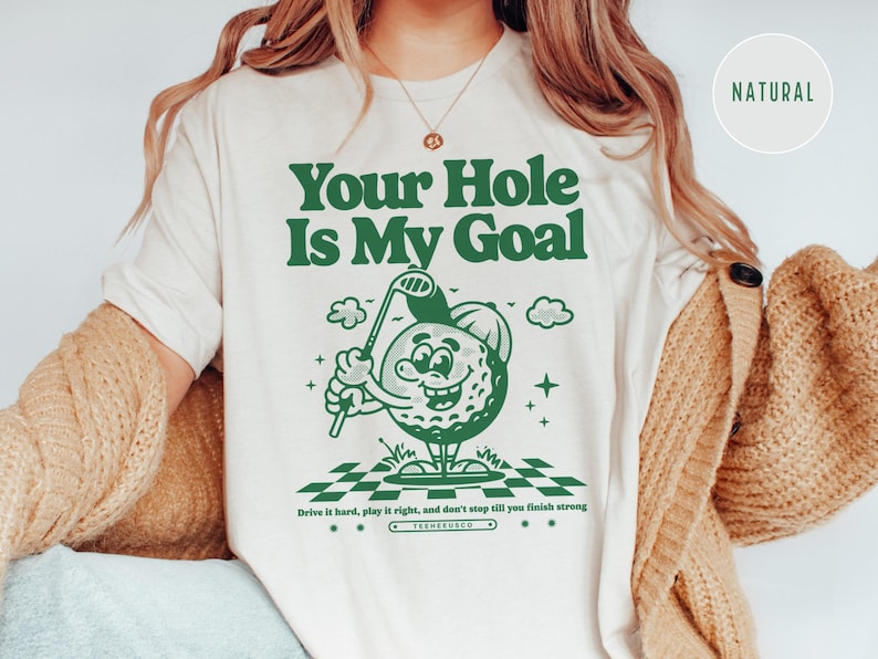Może przedstawiać: Biały t-shirt z zielonym nadrukiem przedstawiającym kresk&oacute;wkową piłkę golfową trzymającą kij golfowy. Tekst "Your Hole Is My Goal" jest nadrukowany na zielono nad grafiką. Tekst "Drive it hard, play it right, and don't stop till you finish strong" jest nadrukowany na zielono pod grafiką.