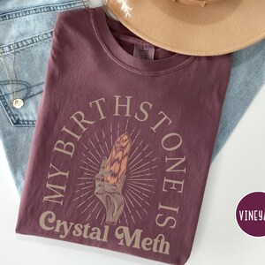 May include: A maroon t-shirt with the text "My Birthstone Is Crystal Meth" in a light brown font. The text is surrounded by a circle with a stylized sunburst design and a hand holding a crystal.