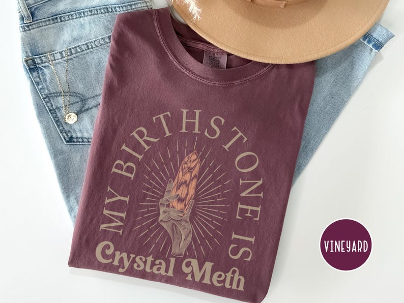 May include: A maroon t-shirt with the text "My Birthstone Is Crystal Meth" in a light brown font. The text is surrounded by a circle with a stylized sunburst design and a hand holding a crystal.