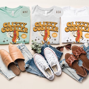 K&ouml;nnte beinhalten: Drei T-Shirts in Mint, Aschgrau und Wei&szlig;, jeweils mit einem Retro-Grafikdesign eines Hotdog-Charakters und dem Text "Glizzy Gobbler". Das Bild enth&auml;lt auch Jeans, Schuhe und Sonnenbrillen, was eine l&auml;ssige &Auml;sthetik erzeugt.