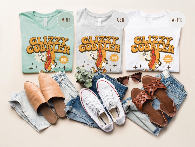 K&ouml;nnte beinhalten: Drei T-Shirts in Mint, Aschgrau und Wei&szlig;, jeweils mit einem Retro-Grafikdesign eines Hotdog-Charakters und dem Text "Glizzy Gobbler". Das Bild enth&auml;lt auch Jeans, Schuhe und Sonnenbrillen, was eine l&auml;ssige &Auml;sthetik erzeugt.