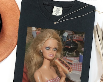 May include: A black crew neck t-shirt featuring a graphic print of a blonde doll with a disgusted facial expression holding a hot dog. The shirt has a Comfort Colors brand label at the inner neckline and a thin gold-tone necklace resting on top.